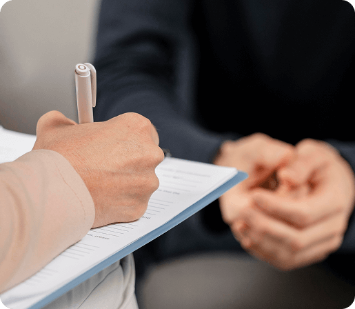 She Knows Alt Text said: Close-up image of a therapy or counseling session, showing a therapist taking notes on a clipboard while the client sits across with hands clasped. The focus is on the interaction, highlighting a professional and empathetic setting.