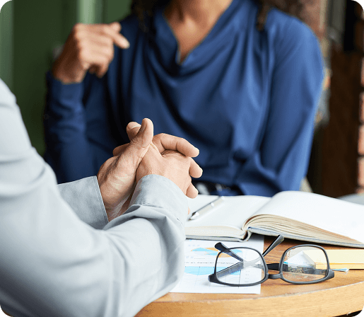 Two people are seated at a table engaged in a serious conversation, with one person's hands clasped and a notebook, pen, and eyeglasses on the table. The focus is on the interaction and body language, suggesting a professional or personal discussion.
